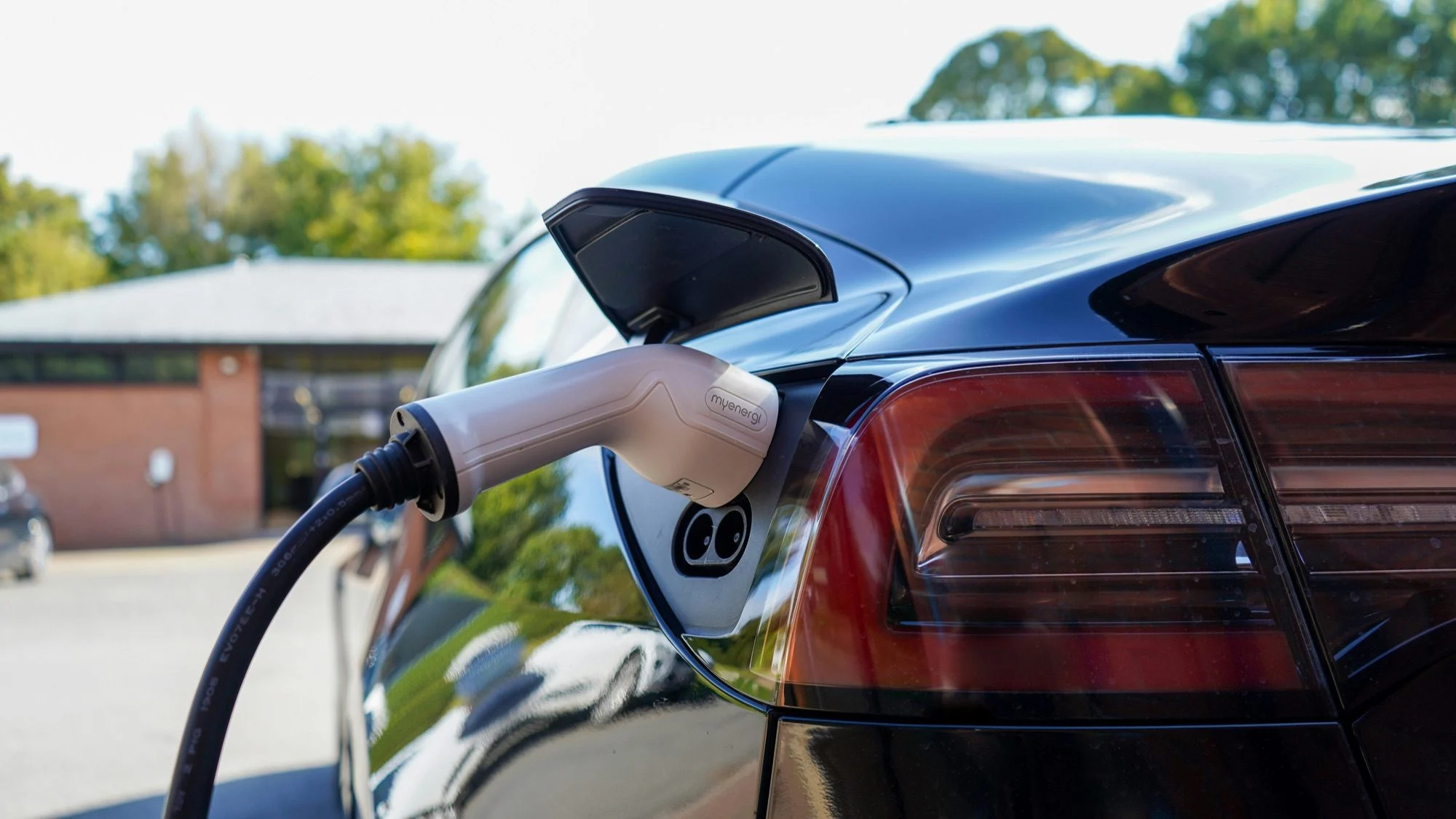 Black Tesla Model 3 plugged into a myenergi home charger, with a red-brick building and trees behind