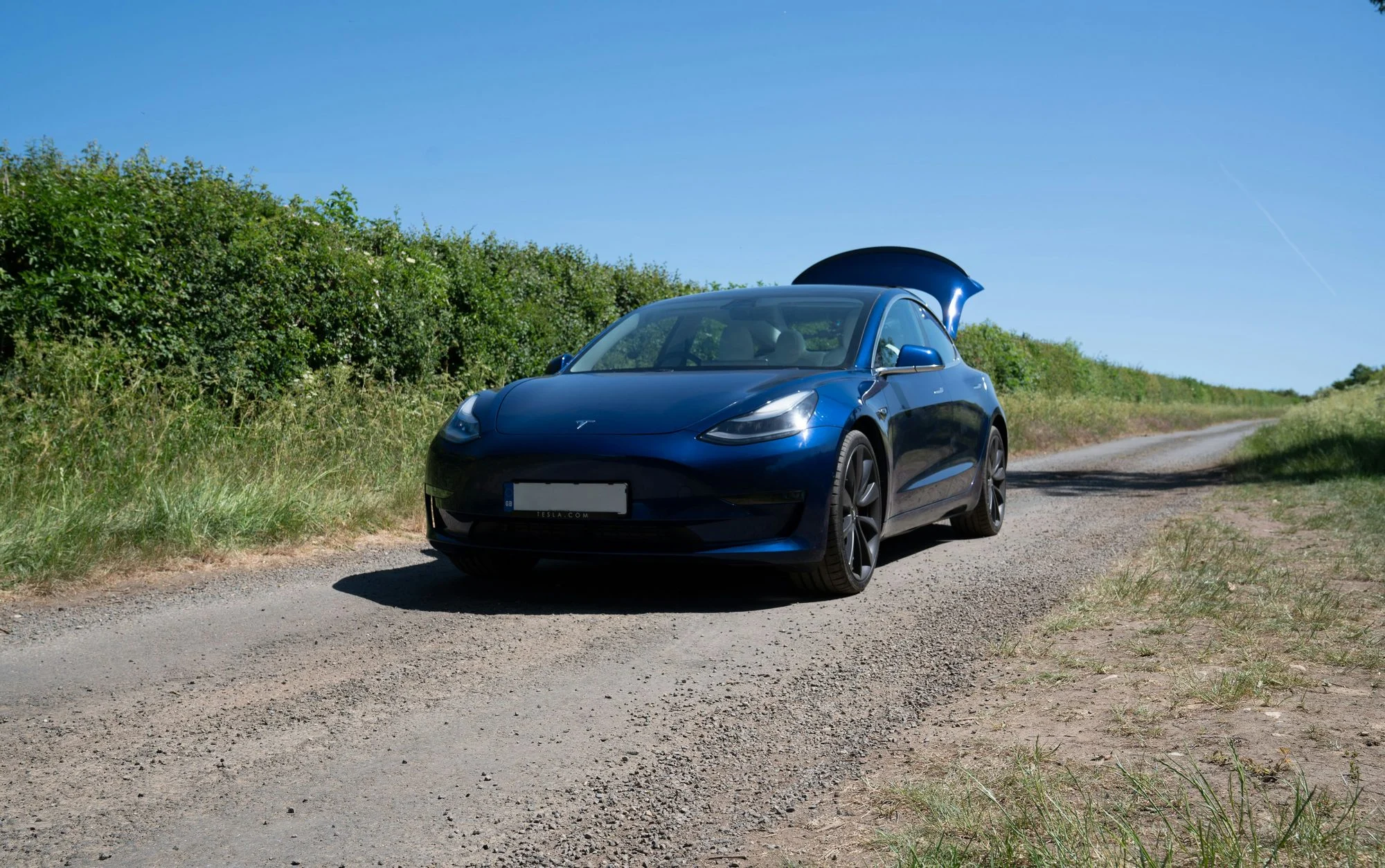 Blue Tesla Model 3 with its boot open on a gravel farm lane between tall green hedgerows on a sunny day