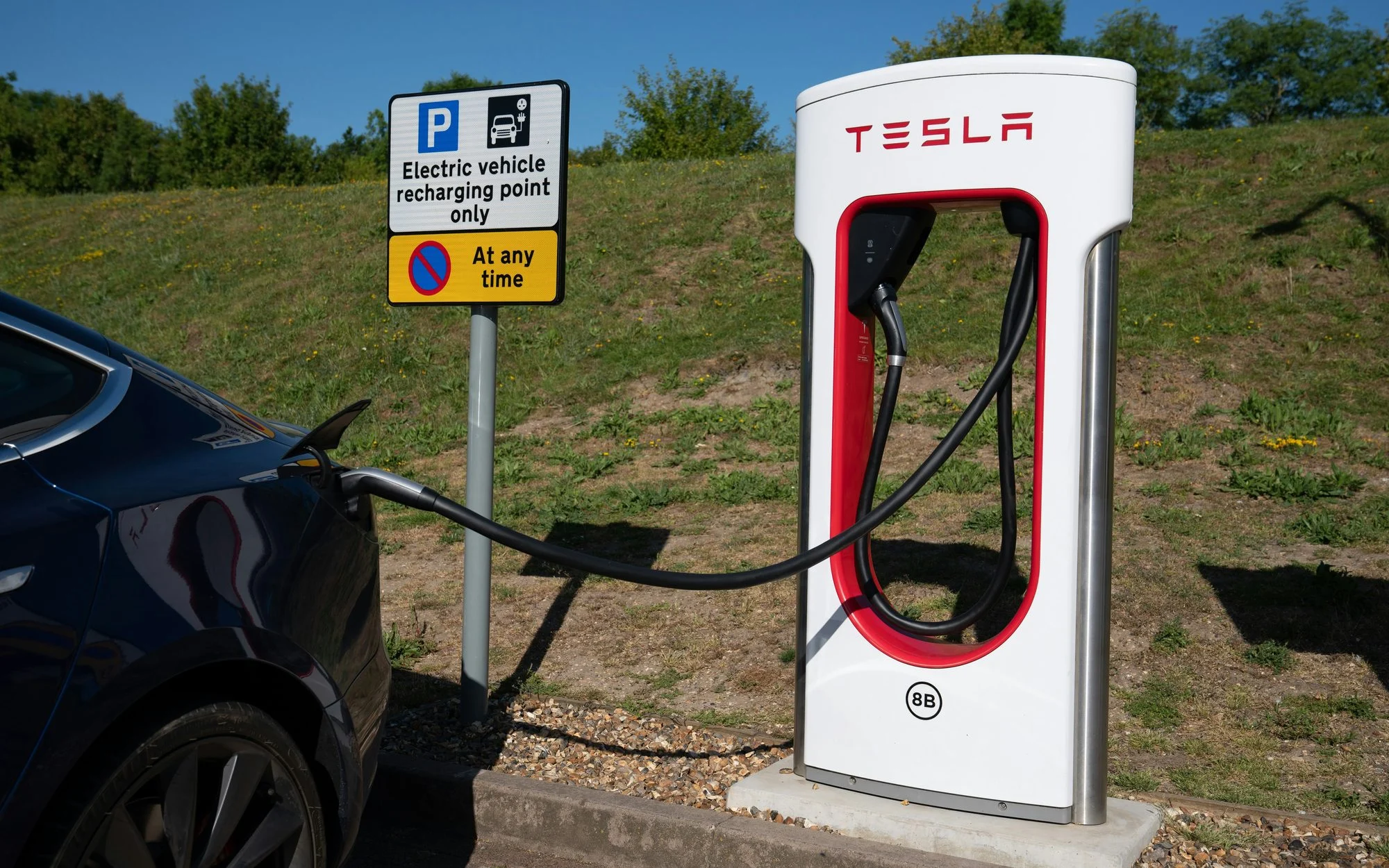 Dark blue Tesla Model 3 plugged into a Tesla Supercharger stall beside a UK sign reading Electric vehicle recharging point only