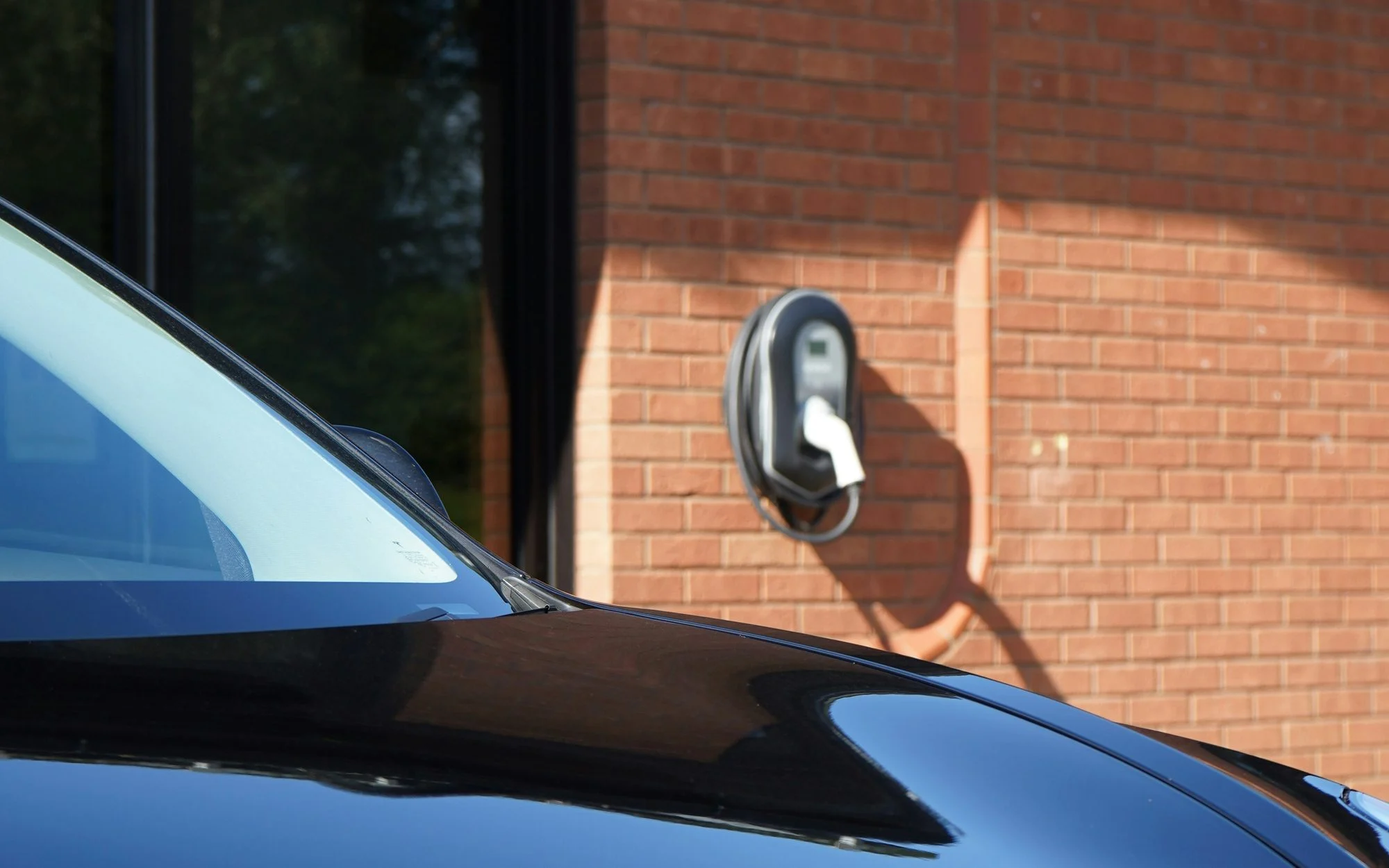 Bonnet and windscreen of a dark blue Tesla Model 3 parked beside a home charger on a red-brick wall